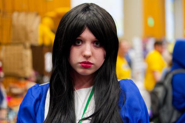 Woman in blue dress and overly perfect black hair looks foreward with a blank facial expression in what seems to be a yellow ballroom with yellow-shirted people in it.