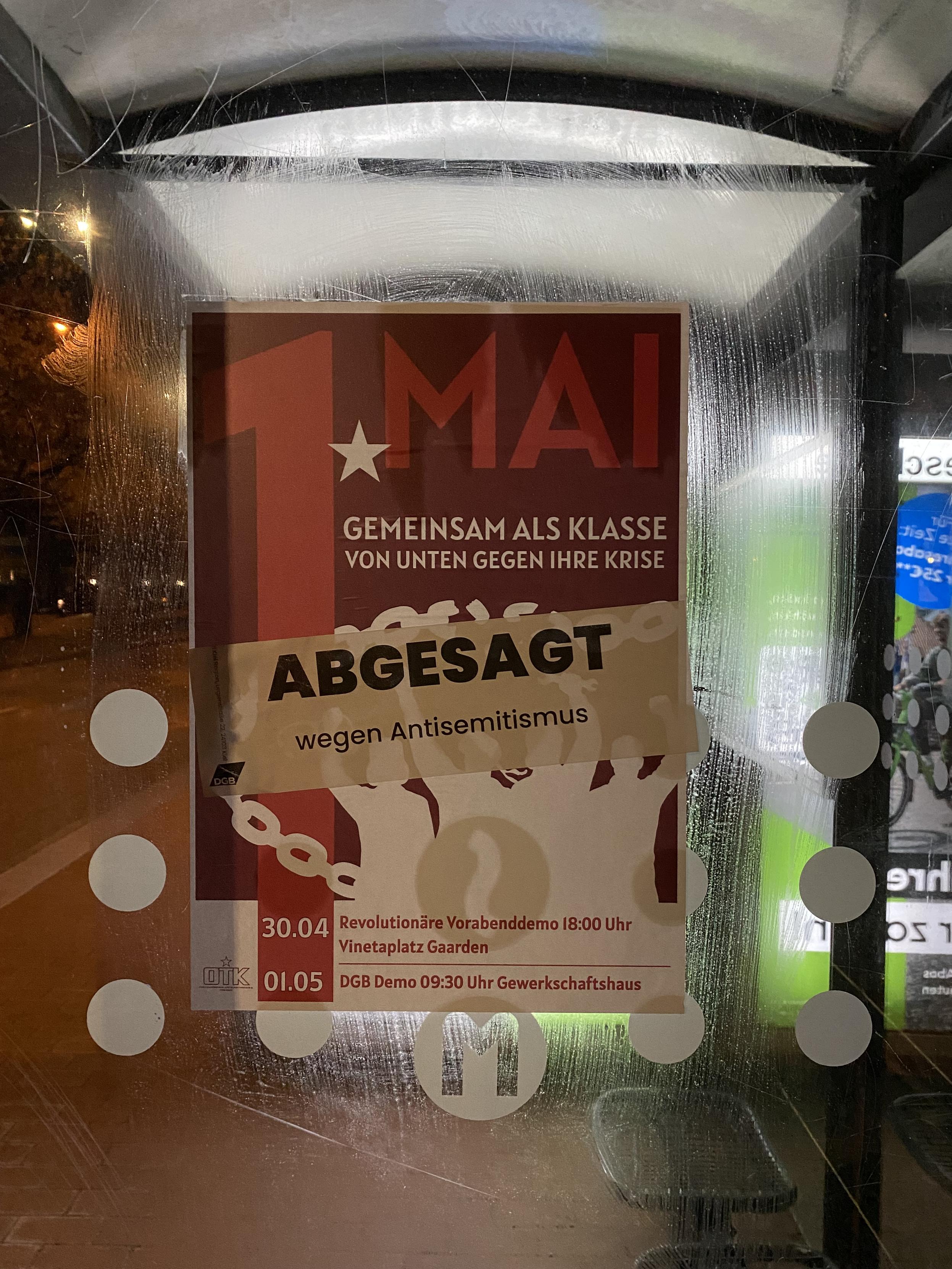 Plakat für Demos zum ersten Mai von autoritärer Gruppe. Darüber klebt „abgesagt wegen Antisemitismus“. 

Das Plakat hängt an einer Bushaltestelle. Die Demo und die VorabendDemo steht unter dem Motto „gemeinsam als Klasse von unten gegen ihre Krise“. Unten in sehr klein Ort und Zeit und daneben steht die organisierende Gruppe. Das Bild scheint in der Nacht aufgenommen.