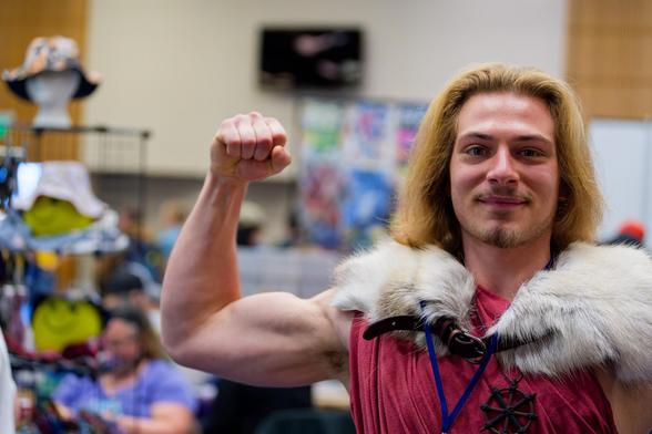 Man defies stereotype by flexing a bicep as a "Fighter" character in the multiverse that includes dungeons and dragons;  he exudes lawful good which grates just a touch for the neutral-chaotic foxographer.  he looks awesome with a red shirt, medium length shaped hair and fur around his shoulders and behind that blurred out mechandise and booths like hats and anime characters and banners and stuff