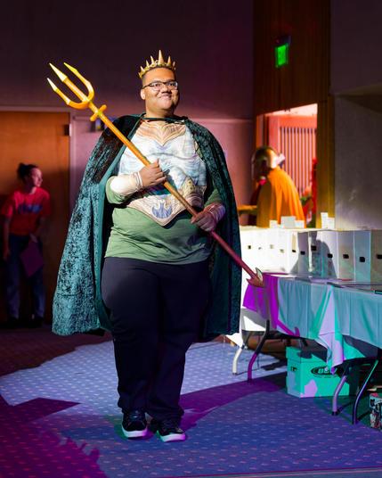 Smiling black man wears green shirt, breastplate and blue cape with many pointed crown and holds trident down runway with EXIT sign and boxes of comics for sale from a vendor behind him and sombody yellow queued up to go next behind him