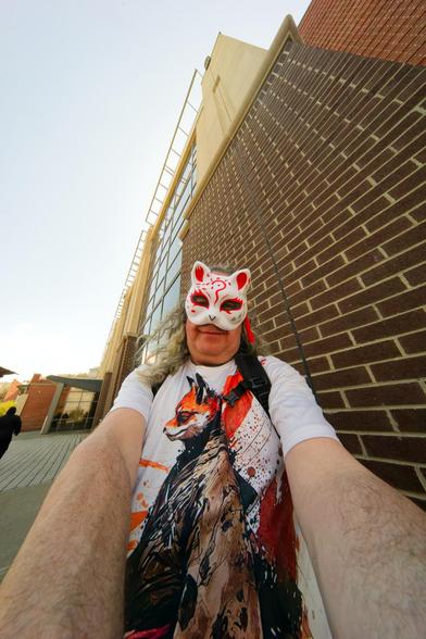 A man's arms are wildly distorted as they approach the bottom corners of the frame,  he's got bare arms under a short sleeved shirt with an orange picture of a red fox furry character dressed like a samurai and he's surely got 8th grader syndrome because he's wearing a white paper mache mask with fox marking despite having copious white hair -- he fills only half the frame and above him looms a brick and (stone?) academic building and gunmetal sky