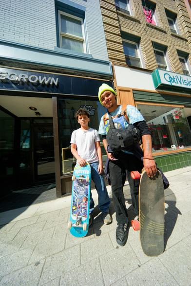 Man and boy pose on street post with skateboards beween skateboard shop and optician