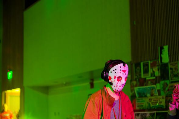 Man in dark orange jacket and blue shirt has white mask with black perforations and orange triangles in front of eerie green lit conference center background