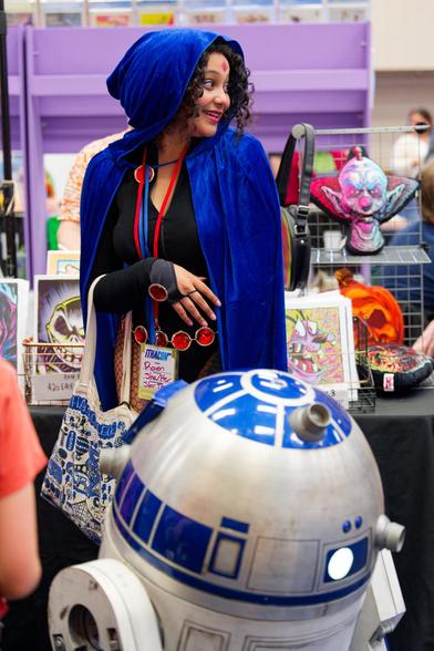 Raven from the Teen titans looks to the left as if she is looking at a friend with R2-D2 in front of her and lots of convention merch behind