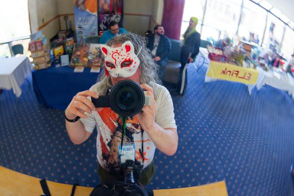 A man with wavy gray heair points fair sized lens at itself reflecting a red and white fox mask, he has a furry samurai fox character on his shirt and a big camera bag in front of him and behind vendor tables with text reflected in a mirror