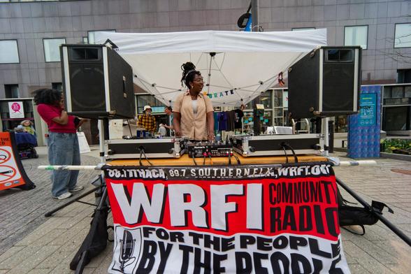 Black woman works control board under tent with a speaker on each side and a sign that say WRFI: Community Radio for the by people by the people
