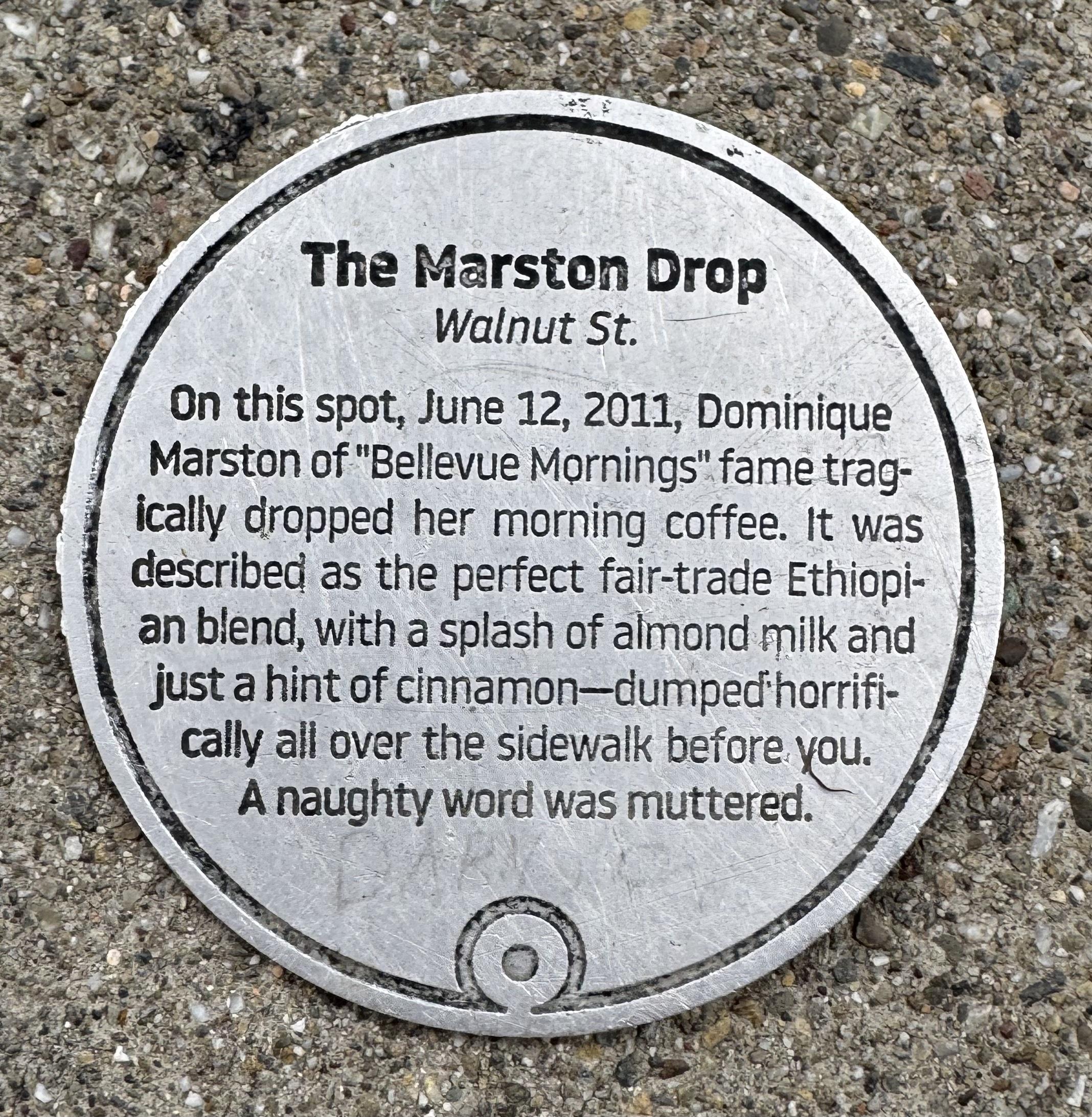 Round metallic disc mounted on the pavement, reading "On this spot, June 12, 2011, Dominique Marston of "Bellevue Mornings" fame tragically dropped her morning coffee. It was described as the perfect fair-trade Ethiopian blend, with a splash of almond milk and just a hint of cinnamon-dumped horrifically all over the sidewalk before you. A naughty word was muttered."