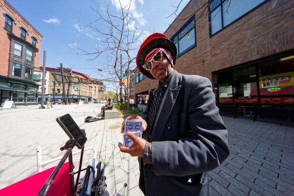 Black man with big red hat and heart-shaped glasses with American flag colors and grey suit shuffles a deck of cards on the east side of the Ithaca Commons