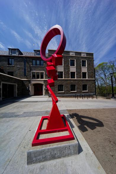 Red metal sculpture with a ring on top casts interesting shadow on concrete and dirt with stone building in background