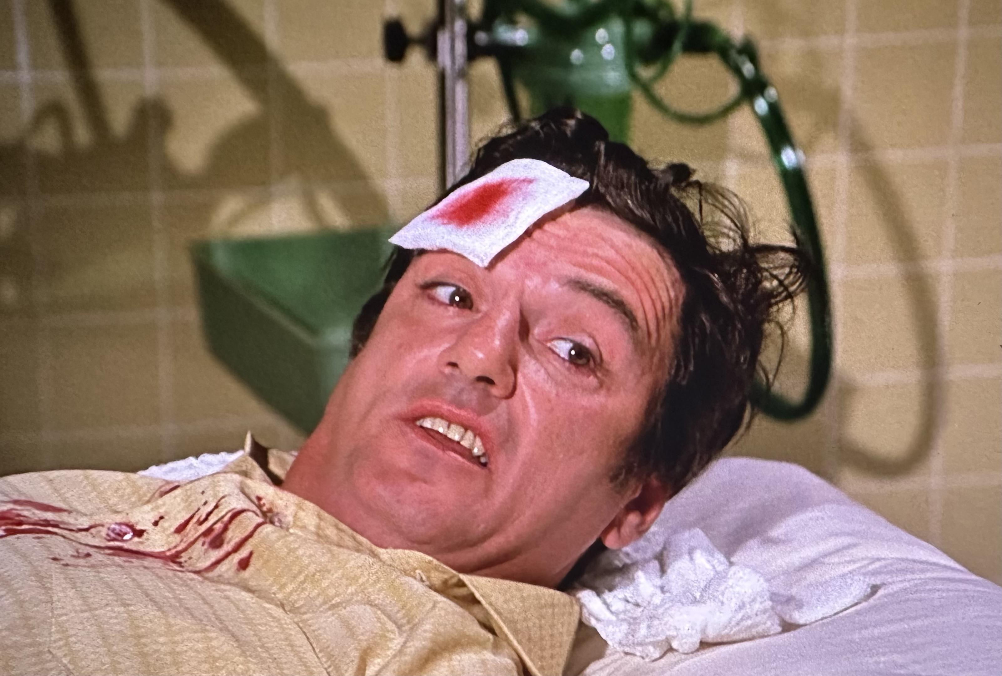 Actor William Campbell playing a drunk driver lies on a hospital bed with a bandage on his head and blood on his shirt.