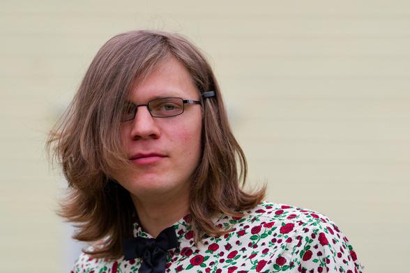 Young man with long straight hair curled up at the ends wears a shirt with little roses printed on it with a creamy yellow background behind