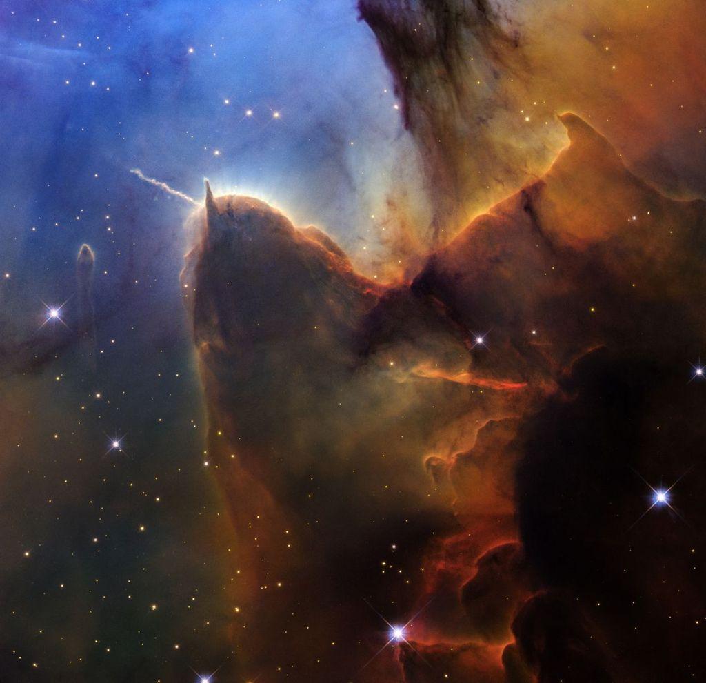 A tightly cropped Hubble view of a vast star-forming region known as the Trifid Nebula. The top left is bright blue. Brown and amber colors run from top right through the center in irregular, overlapping lines to the bottom-center. At bottom right, the view is almost black. Tiny, amber-colored stars appear throughout the scene. Toward the left there is a prominent brown shape that looks like a head with two horns. The left horn points left and is wavy. The right horn is triangular and points up. The brown dust continues, flowing down, as if along a back, and up toward the top right. A prominent line, about the same length as the left horn, appears below the middle of the body, and changes from orange to red. A small, separate semi-transparent pillar is left of the head. A few slightly larger, blue foreground stars with four diffraction spikes appear in the bottom half.