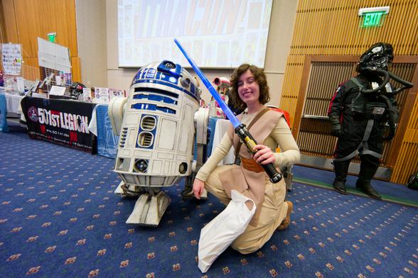 Woman dressed as a star wars princess with blue light saber kneels next to a robot that looks like a trashcan on top of three wheel housings with a rotating dome head on top;  a stormtrooper guards the exist and the table of the 501st legion can be seen in the background