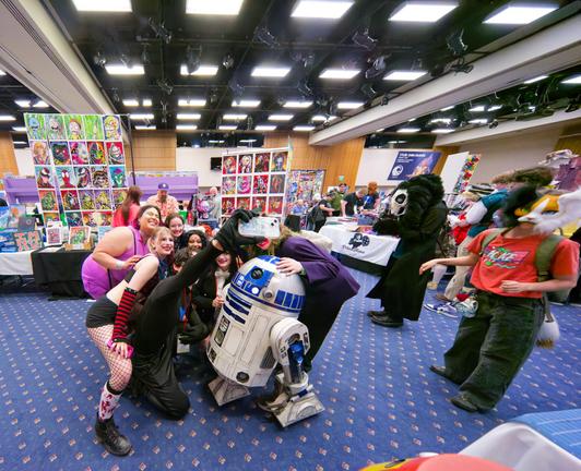 A whole bunch of people pose for a cell phone snap with R2D2 in a wide angle shot that shows a whole lot of color art for sale by bendors and a dramatic array of lights on the ceiling with onlookers wearing maksed costumes looking on from the right side