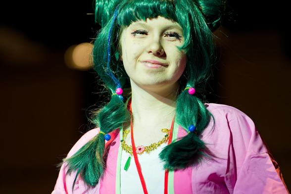 Close shot of woman wearing green wig and white shirt with pink jacket, pink beads in hair and pink lipstick