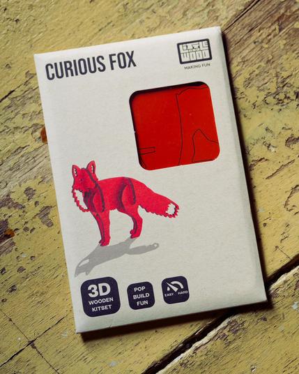 Beige rectangular package roughly 4x6 labeled CURIOUS FOX with a window showing orange precut cardboard and a 3-d render of a red fox made from interlocked flat pieces casting a shadow on a yellow diagonally oriented wood floor