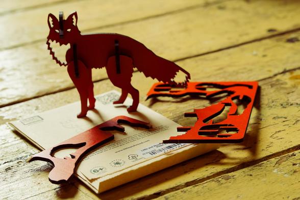 Three dimensional fox model glare at me with eyes lit by the yellow floor behind.  He stands on the beige package that he came in and the piece of cardboard I pressed pieces out to make him with