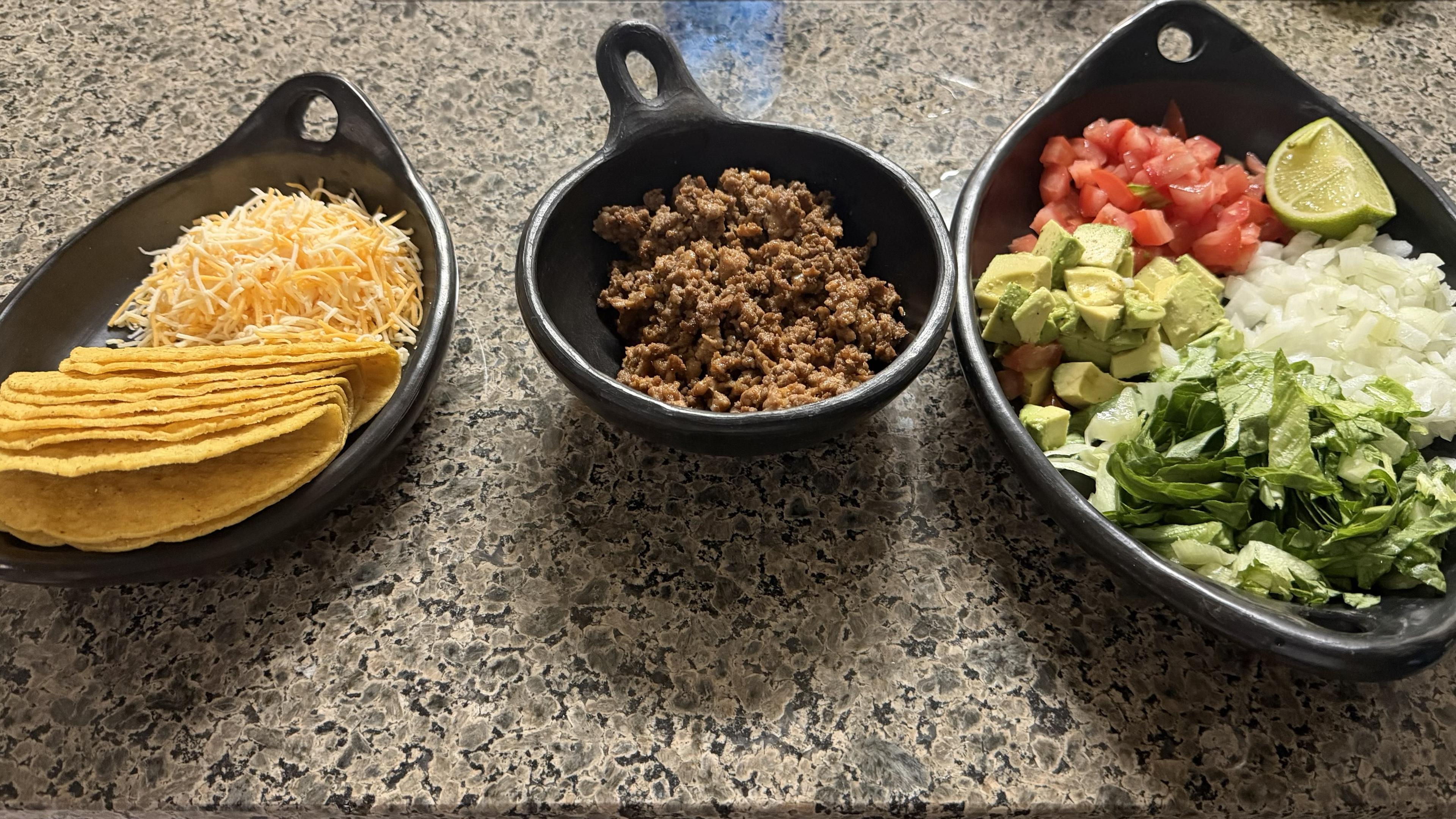 Picture of all the fixings for hardshell tacos, including lettuce, tomato, onion, cheese, and meat all neatly displayed in three clay bowls