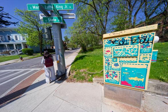 In the 1/3 right find a yellow and blue painted electric box with comic-book like panels depicting swa scenes and a grafitti tag and behind it a grass lawn, leafing out trees and to the left and intersection with signs and a person standing with their back to me.