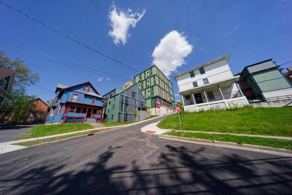at the bottom a road twists and vanishes towards the center with distorted builindgs in bold colors look tiny under a mostly white sky with maybe three cumulus clouds