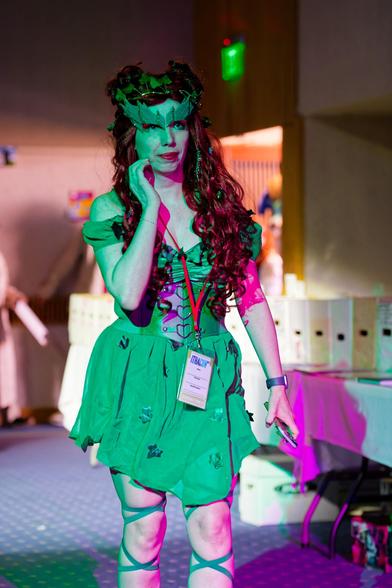 Woman with green dress with green waist cincher cinched with hearts and green mask and long red drills to her waist has laces crossing across legs