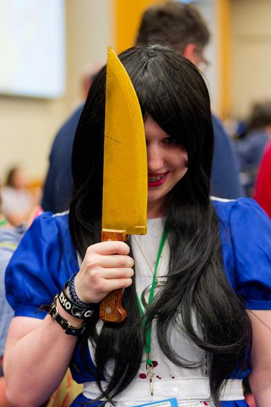 Girl in blue and white dress covers one eye with a huge yellow knife while she flashes an evil smile