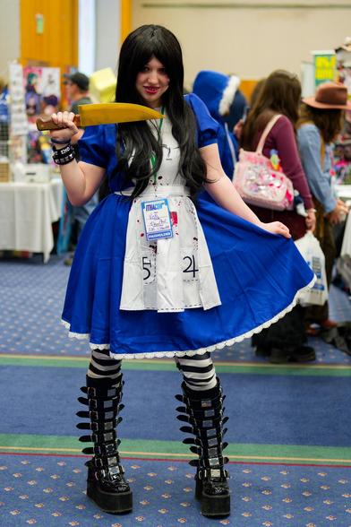 Whole body view of evil alice showing her tall boots with outrageous buckles and black and white striped leggings under a knee lenght blue dress with white hem and a sort of bib in front with astrological symbols with an unfortunately congested background