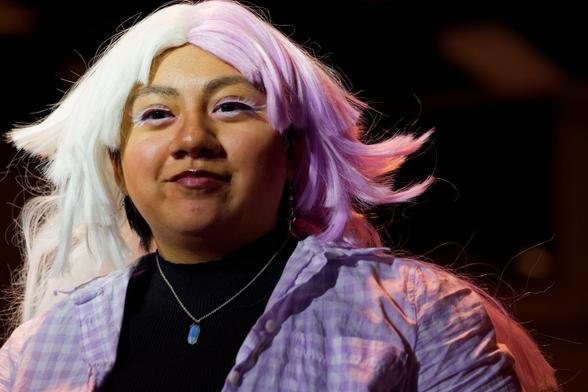 Woman wears a white wig with hair that curls up to the sides and has a dark shirt with a checked shirt over it