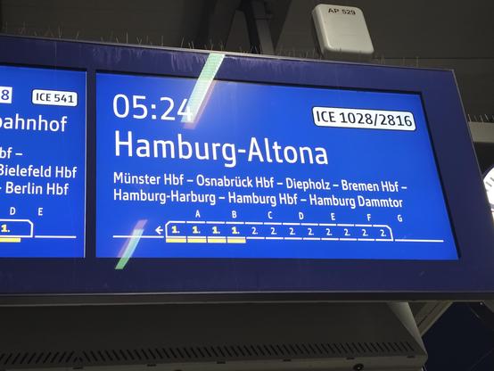 Ein digitales Informationsdisplay mit blauem Hintergrund und weißem Text zeigt Zugverbindungen an. Auf dem Hauptbildschirm stehen die Zeit „05:24“, die Zugnummer „ICE 1028/2816“ und das Ziel „Hamburg-Altona“. Die angegebene Route lautet „Münster Hbf – Osnabrück Hbf – Diepholz – Bremen Hbf – Hamburg-Harburg – Hamburg Hbf – Hamburg Dammtor“. 