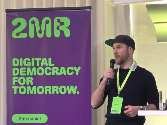 Person, die neben einem großen violetten Banner spricht. Das violette Banner auf der linken Seite enthält den Text „2MR“ in großen grünen Buchstaben, gefolgt von „DIGITAL DEMOCRACY FOR TOMORROW.“ und unten in einem grünen Feld „2mr.social“. Auf der rechten Seite hält eine Person mit Bart ein schwarzes Mikrofon an den Mund. Die Person trägt eine dunkle Baseballkappe, einen dunklen Pullover und ein hellgrünes Schlüs Schlüsselband mit einem kleinen weißen Namensschild, auf dem „2MR“ steht