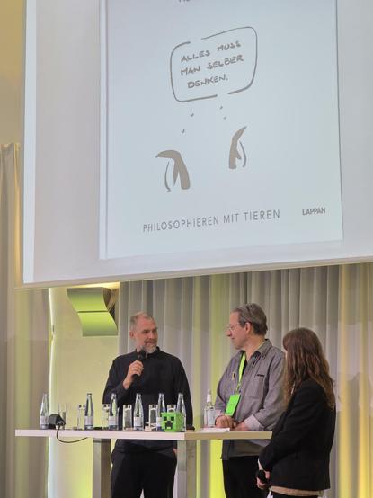 3 Personen stehen hinter einem weißen Tisch vor einer großen Projektionsfläche. Die Leinwand zeigt oben den Text „ALLES MUSS MAN SELBER DENKEN.“, darunter zwei einfache schwarze Zeichnungen sowie unten die Wörte Wörter „PHILOSOPHIEREN MIT TIEREN“ und „LAPPAN“. Die Person links hält ein Mikrofon und trägt ein dunkles Oberteil, die mittlere Person trägt eine Brille und ein helles Hemd mit einem grünen Band, und die dritte Person ist von hinten in dunkler Kleidung zu sehen. 
