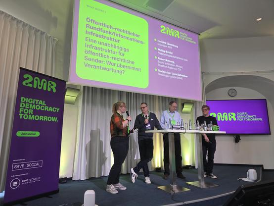 4 Personen stehen auf einer Bühne vor einer großen Projektionsleinwand und einem vertikalen Banner. Eine Person auf der linken Seite hält ein Mikrofon, zwei Personen stehen hinter einem hohen, schmalen Tisch mit Wasserflaschen und eine vierte Person steht am rechten Ende des Tisches. Das vertikale Banner links zeigt den Text „2MR DIGITAL DEMOCRACY FOR TOMORROW. 2mr.social veranstaltet von SAVE SOCIAL in Zusammenarbeit mit KING | CONSULT KOMMUNIKATION“. 

Die große Leinwand im Hintergrund z…