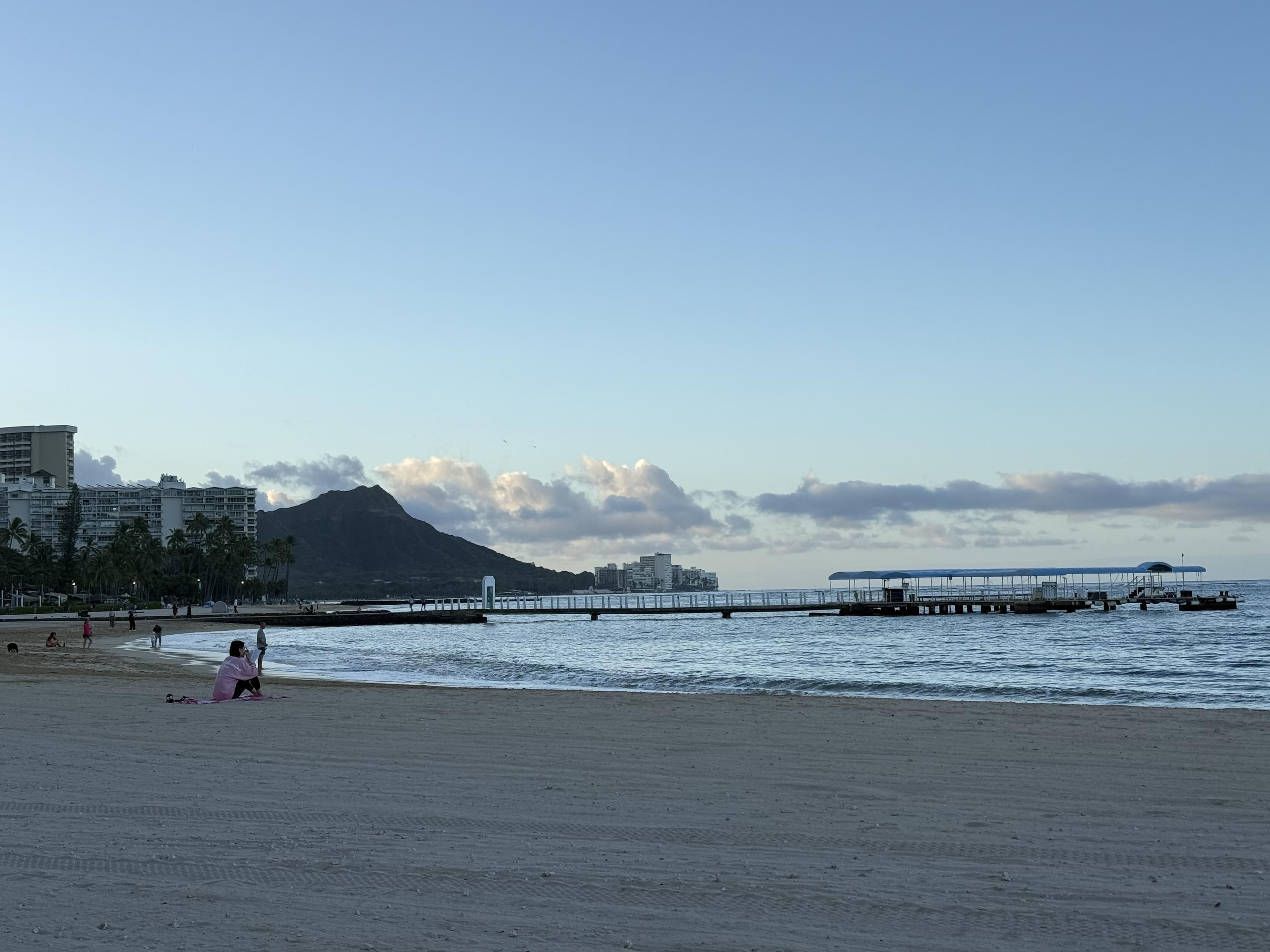 A serene beach scene featuring gentle waves lapping at the shoreline. A mountainous backdrop is visible, along with a long pier extending into the water. Several people can be seen enjoying the beach, with some seated on the sand. The sky is clear.