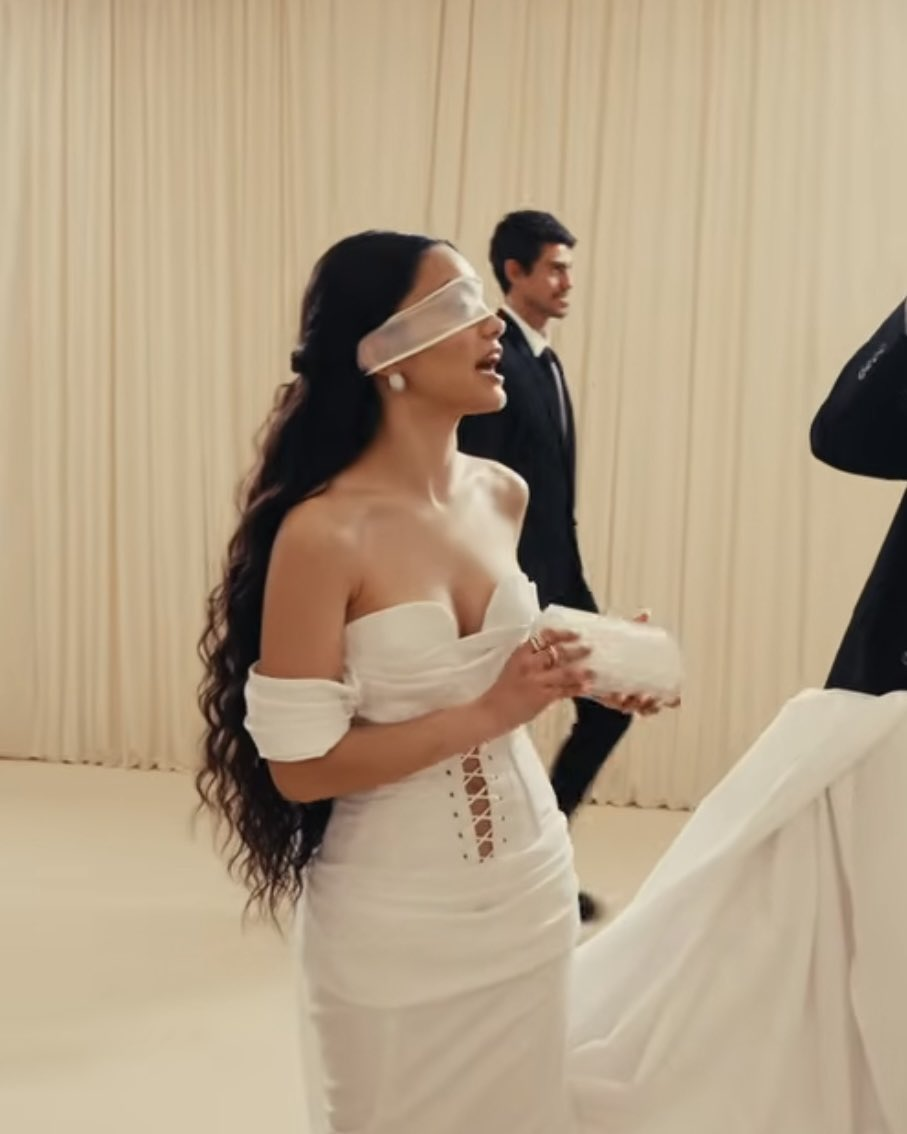 A woman with long, wavy dark hair stands in profile against a backdrop of cream-colored pleated curtains. She wears a white off-the-shoulder gown with a lace-up corset bodice and a sheer white blindfold over her eyes. She holds a small, clear rectangular clutch bag. In the blurred background, a man in a black suit walks past.