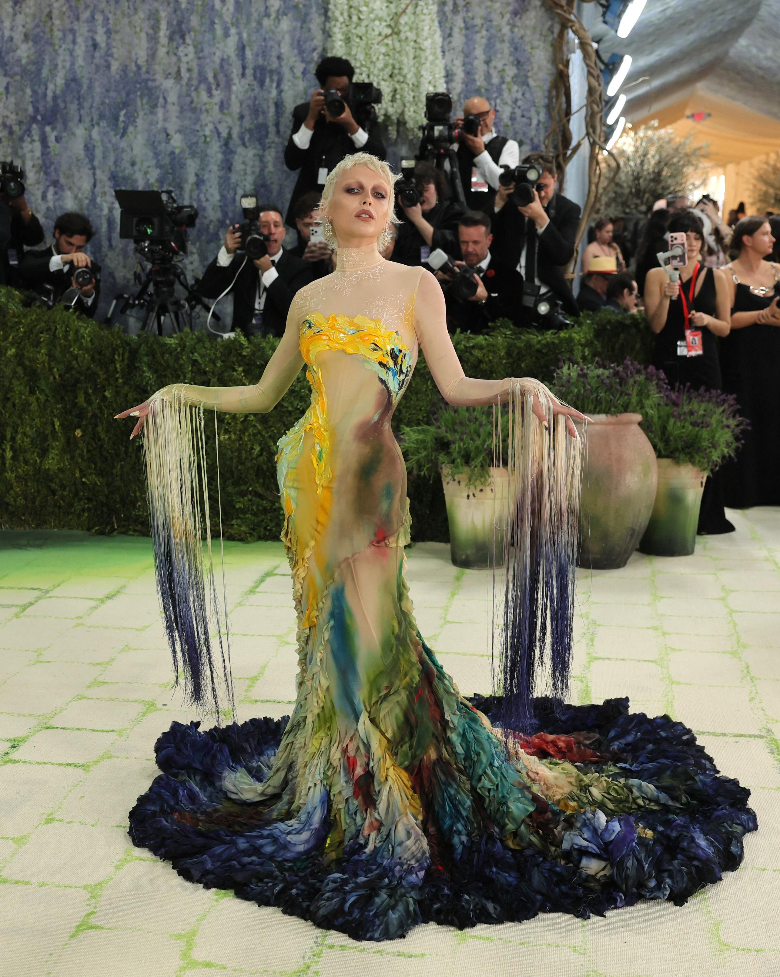 An individual with short blonde hair and dramatic eye makeup stands on a carpeted area, wearing a form-fitting gown with a long train. The dress features a watercolor-like palette of yellow, green, brown, and blue, with long fringe sleeves that transition from light blonde to dark blue. Photographers and onlookers are visible in the background against a floral and greenery backdrop.