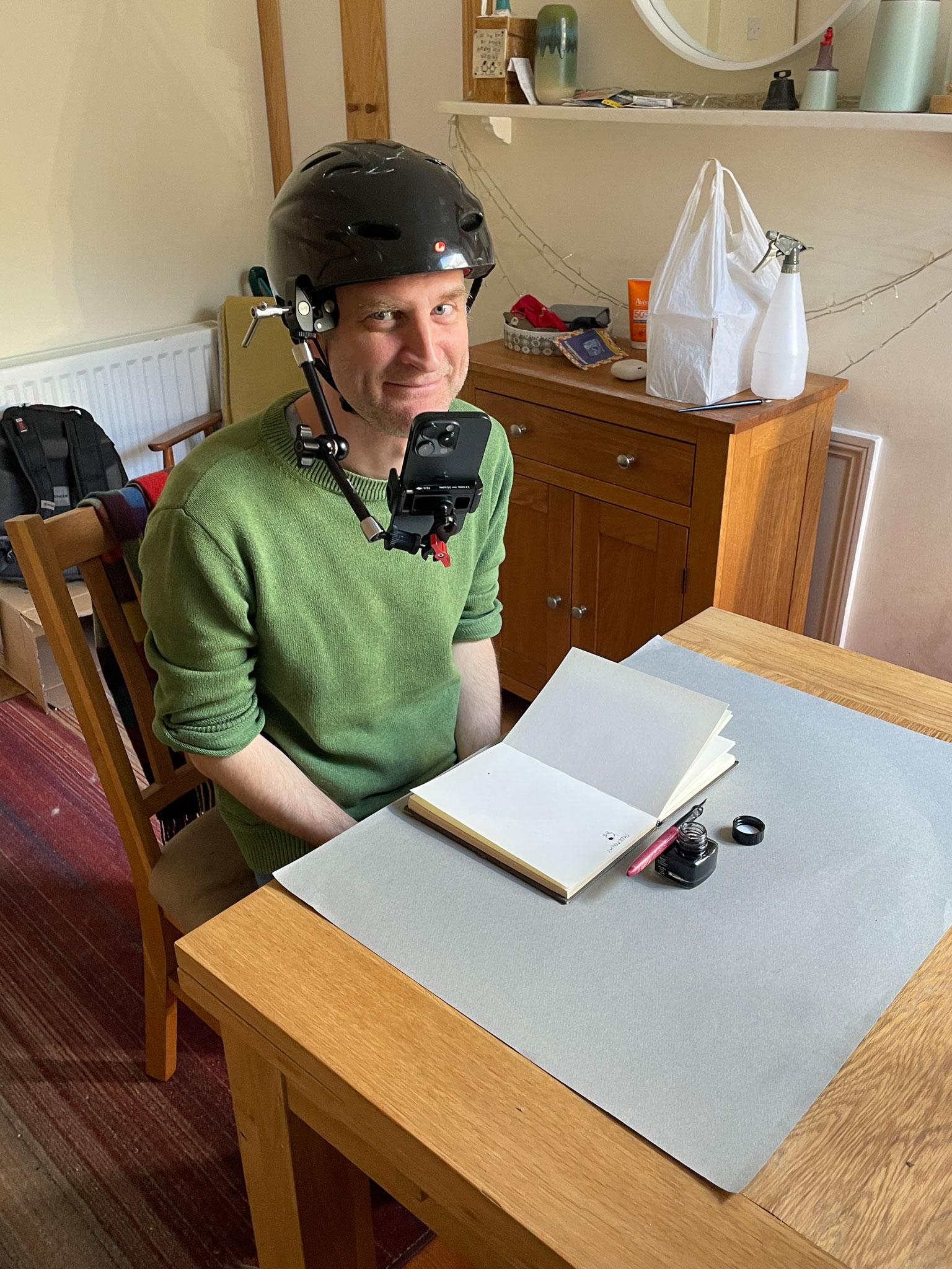 A person with a bike helmet and phone attached to it. Sitting at a table with a sketchbook, ink bottle and ink pen in front of them