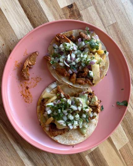 Two delicious looking street tacos with onions and other condiments and diced meat on little tortillas on a purple plate on a wood table.