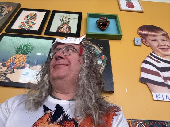 Man with white shirt with a red fox on it has long curly grey hair and has one of those white fox masks with red markings in a little red tassel on his head above his hexagonal glasses and behind him a yellow wall with mostly pictures of pineapples on it (really!)