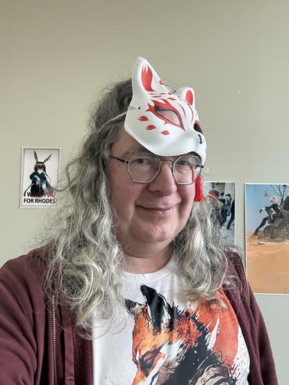 Man with fox shirt and mauve jacket has long drill silver hair with a fox mask on his forehead above octagonal glasses,  to his left is a picture of Amiya from Arknights saying "I WANT YOU FOR RHODES" like Uncle sam and to his left are posters showing his beautiful wife and 2B from Neir Automata.