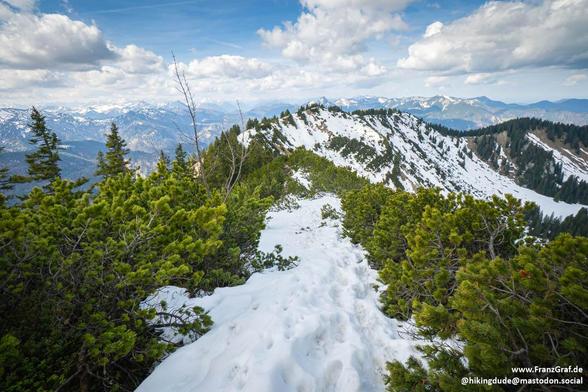 This image captures a stunning and dynamic alpine landscape, showcasing the transition between seasons in a mountainous region. The foreground features a trail blanketed in patches of snow, winding its way through a terrain that is beginning to thaw. The snow's pristine white surface is interspersed with areas of exposed ground, hinting at the arrival of spring.

Flanking the trail are clusters of evergreen trees, their vibrant green foliage providing a striking contrast to the white snow. The …