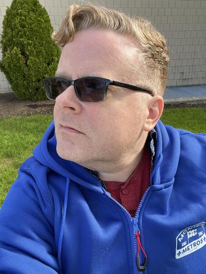 Bill stands outside a building wearing a blue Montreal Metros hooded zip sweatshirt, looking off camera blankly. 
