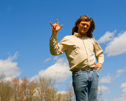 Young man with blue jeans and cream colored buttondown shirt makes hand gesture with pinky, index and thumb extended and others contracted and shoulder length hair;  trees in the background to the lower left and blue sky with cumulus clouds on the other side