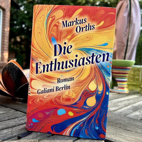 Titelbild des Romans „Die Enthusiasten“ von Markus Orths. Es zeigt einen bunten Strudel, in der Mitte gelb und orange, nach außen hin über rot nach blau übergehend.