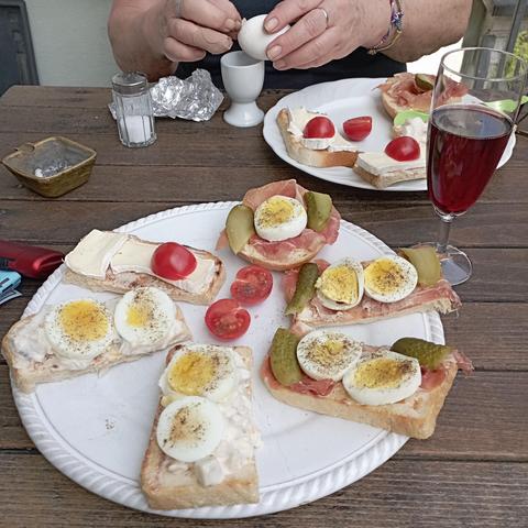 A close-up view of a wooden table featuring plates of open-faced sandwiches and hands peeling a hard-boiled egg. In the upper center, hands are peeling the shell from a hard-boiled egg resting in a white egg cup. A large white plate in the foreground holds several sandwiches topped with sliced hard-boiled eggs sprinkled with pepper, cherry tomato halves, ham, and (🇩🇪 Spreelinge®)slices. To the right, a glass is filled with a red sparkling wine, and another plate of sandwiches is visible in the …
