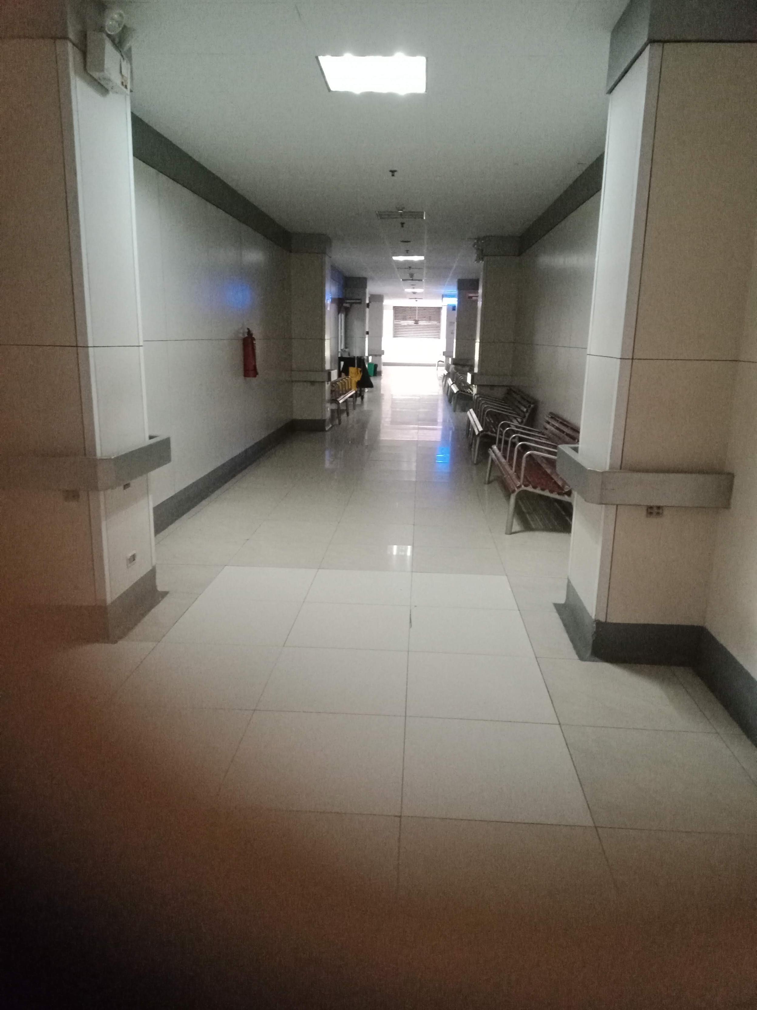 image depicts a long, well-lit hospital hallway with light-colored tile floors and walls, featuring handrails along the sides