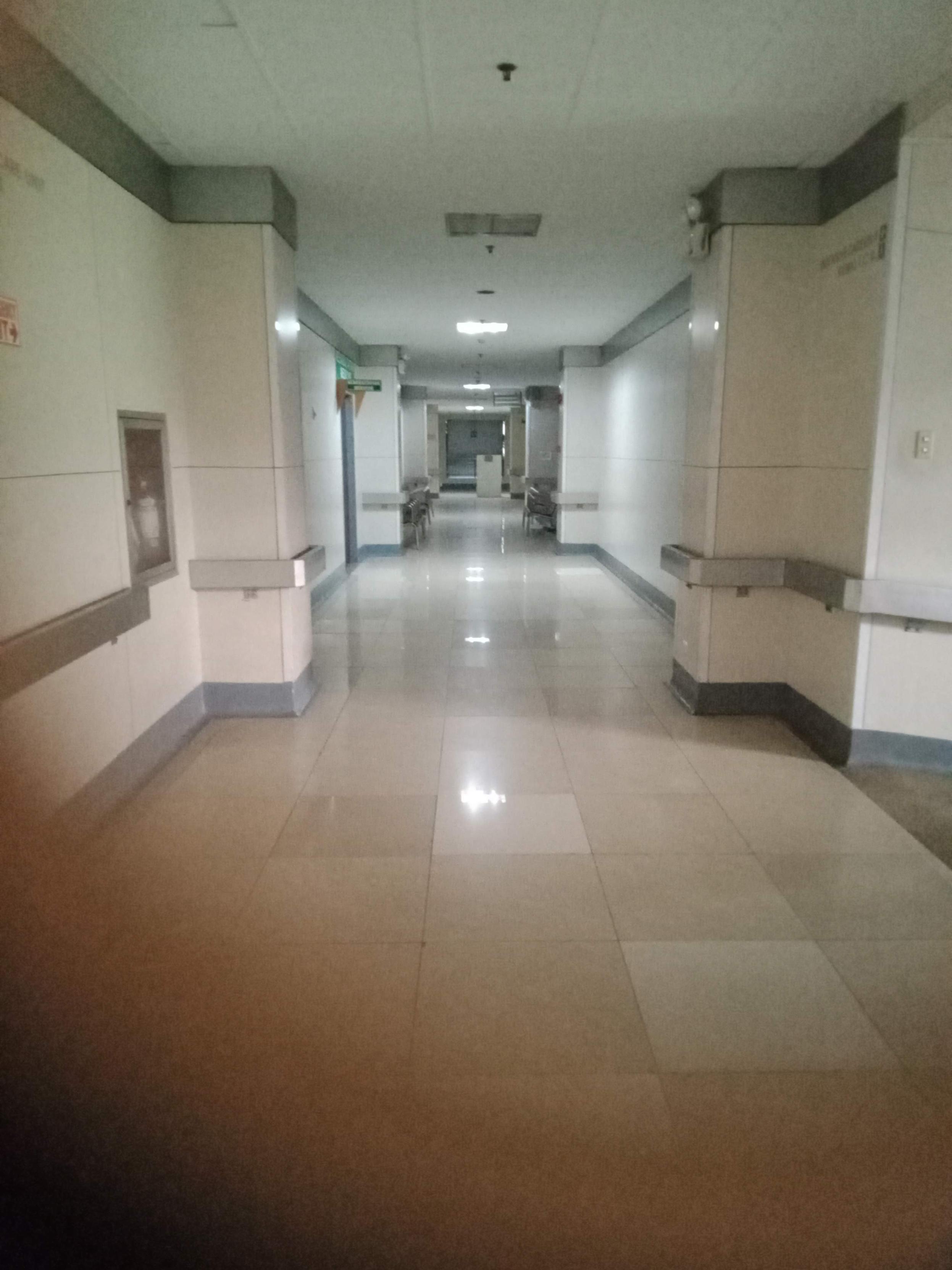 image depicts a long, well-lit hospital hallway with light-colored tile floors and walls, featuring handrails along the sides