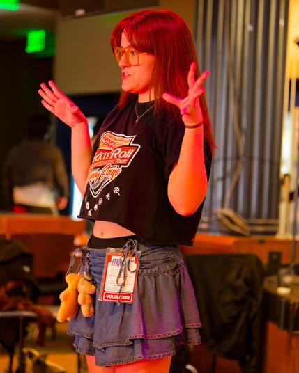 Young woman has denim pleated skirt with a conference lanyard and small stuffed animals and a black t-shirt take leaves an open midriff and auburn hair and big glasses has her hands wide and open sas she talks with a blurry but somewhat confused background
