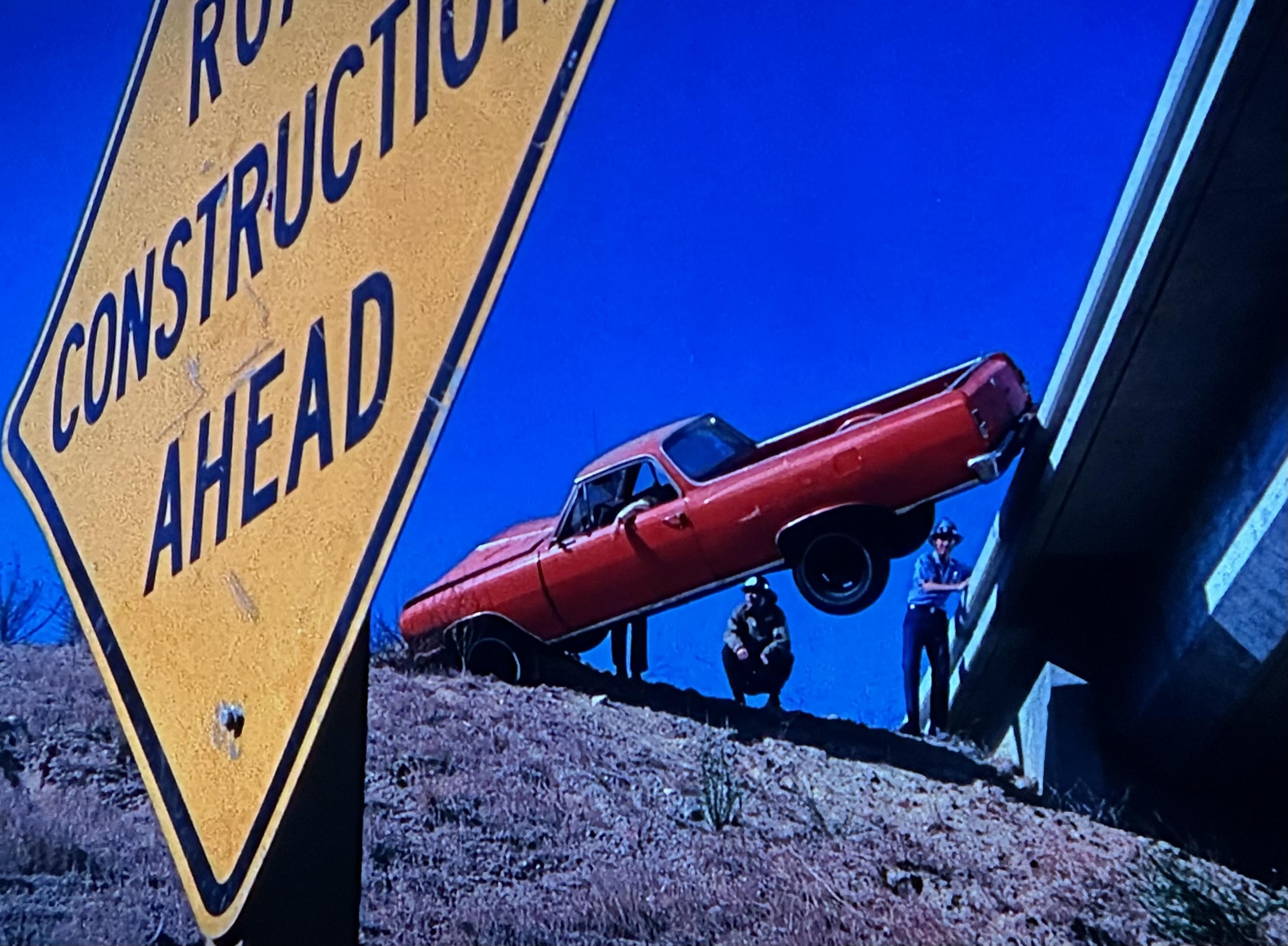 Looking up at an embankment of a California highway overpass where a red truck has gone over the edge and has become wedged between the embankment and the overpass itself. The car is teetering over a very tall gap and a man’s arm is hanging out the window. The firefighters are examining the situation and figuring out what to do next.