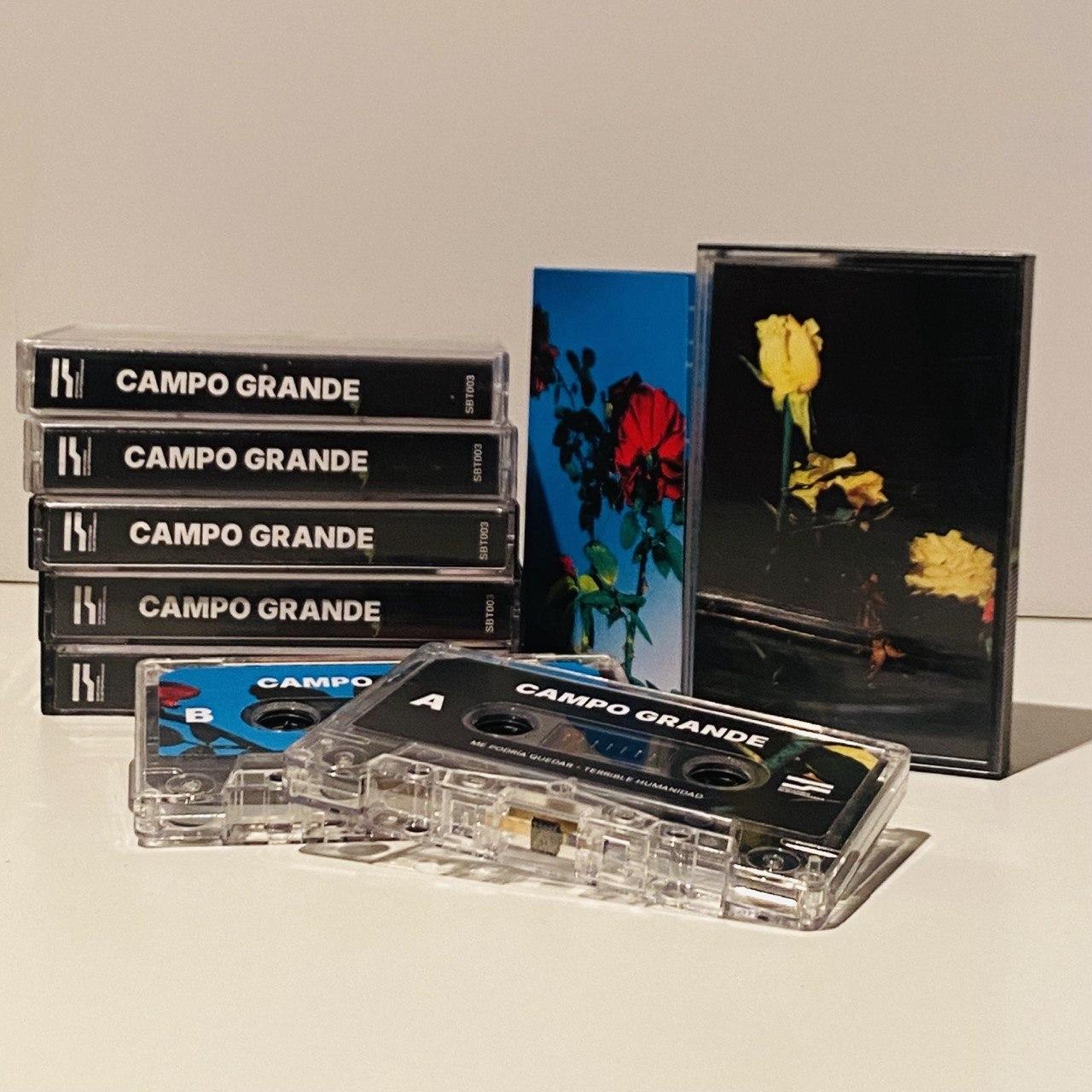Cassettes de Campo Grande. En el fondo hay varios en las cajas puestos uno encima de otro a la izquierda. A la derecha se ve la portada y el interior del libreto. La portada tiene un fondo negro con unas rosas amarillas, el interior en contraste un fondo azul con unas rosas rojas. Estas mismas imágenes se usan como etiquetas de la cara A y B en los cassettes que, a modo de muestra, hay puestos dos fuera de las cajitas en el frente.