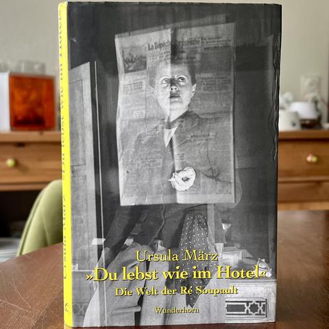 Titel des Buches "Du lebst wie im Hotel – Die Welt der Ré Soupault" von Ursula März. Es zeigt eine Schwarzweiß-Fotografie von Ré Soupault, die in Hose und Sakko so an einen Tisch gelehnt ist, dass es leicht wie sitzen aussieht. Sie hält eine fast abgebrannte Zigarette in ihrer linken Hand. Mit der rechten Hand stützt sie sich am Tisch ab.